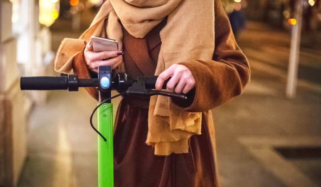Una ragazza con sciarpa e cappotto tiene una mano sul manubrio di un monopattino elettrico mentre nell'altra mano stringe lo smartphone