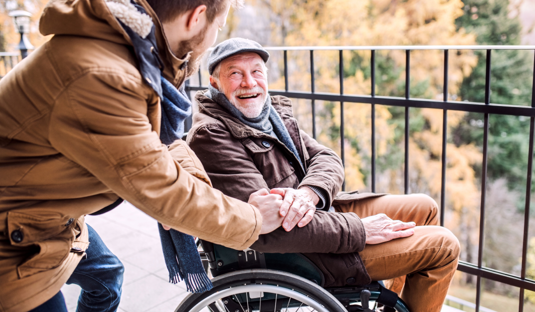 Un anziano in sedia a rotelle all'aperto ride tenendo la mano di un uomo più giovane che lo accompagna
