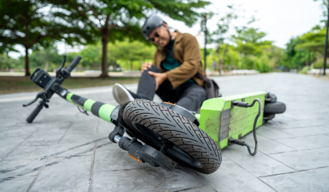 Un uomo con casco e occhiali da sole si tocca il ginocchio dolorante dopo essere caduto da un monopattino